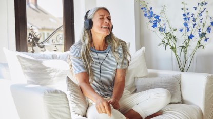 A mature woman in a striped shirt sitting on a white couch while wearing headphones to tap into the benefits of neural nostalgia